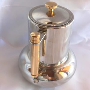 Può includere: Una caffettiera in metallo argentato lucido con corpo cilindrico e base arrotondata. È dotata di un manico verticale color oro e di un pomello coordinato sul coperchio incernierato. La superficie è altamente riflettente con riflessi metallici.