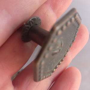 May include: A bronze-toned metal cabinet knob featuring a hexagonal face with a raised beaded border and a central embossed design. The knob includes a cylindrical stem and a circular base plate with a decorative scalloped edge finish.