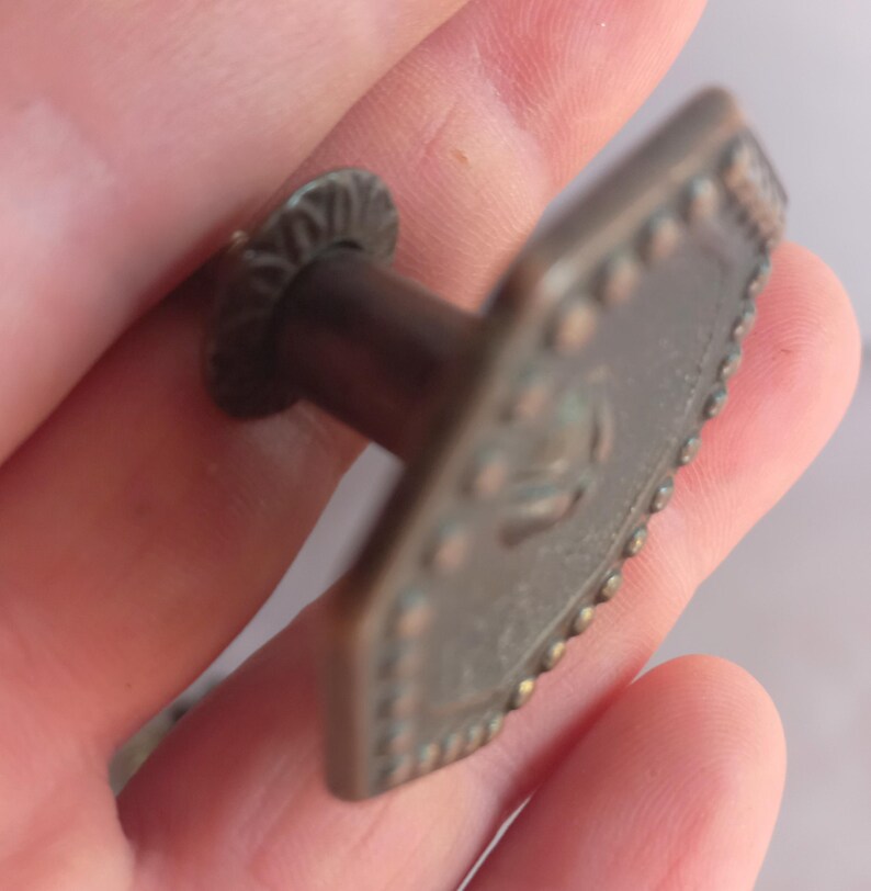 May include: A bronze-toned metal cabinet knob featuring a hexagonal face with a raised beaded border and a central embossed design. The knob includes a cylindrical stem and a circular base plate with a decorative scalloped edge finish.