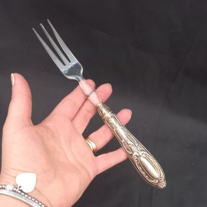Op de afbeelding: Zilveren vork met drie tanden en een decoratief handvat. Het handvat heeft een sierlijk ontwerp met een helder, cilindrisch gedeelte dat de tanden van de vork verbindt. De vork wordt in een hand gehouden tegen een donkere achtergrond.