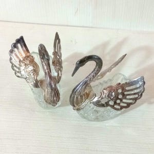 May include: Two decorative swan-shaped glass and silver-tone metal containers. The swans have outstretched wings and curved necks. The containers are clear glass, and the metal has an ornate design.