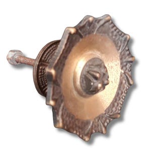 Puede incluir: Pomo de armario en tonos bronce con una cara decorativa en forma de estrella. El pomo presenta un diseño central circular con un patrón texturizado en relieve y un perno roscado para la fijación. El diseño del pomo tiene una estética antigua o vintage.