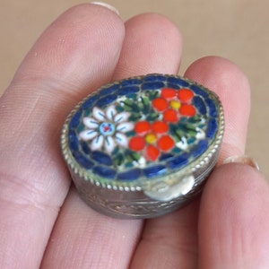 May include: Oval-shaped mosaic trinket box with a silver-colored metal frame. The lid features a floral design with white, red, and blue mosaic tiles. The box is held in a hand.