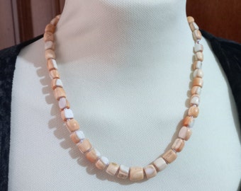 May include: A necklace featuring square, peach-toned beads with white highlights. The beads are strung together, creating a continuous loop. The necklace is displayed on a white mannequin bust against a dark backdrop.