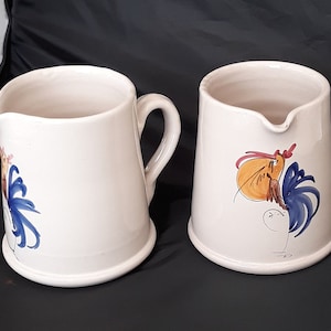 May include: Two white ceramic jugs with rooster designs. One jug has a blue rooster with a yellow beak and red comb. The other jug has a blue and yellow rooster with a red comb.