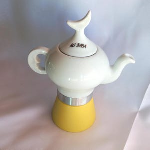 May include: A white ceramic teapot with a yellow base and a silver band. The lid features a curved handle and the text "ALI BABA". The teapot has a curved spout and a rounded handle.