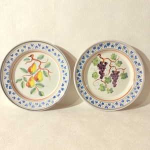 Peut inclure: Deux assiettes décoratives en céramique. L'une représente une branche de poires avec des poires jaunes et oranges et des feuilles vertes. L'autre présente une vigne avec des raisins violets et des feuilles vertes. Les deux assiettes ont un motif de cœurs bleus sur le bord.