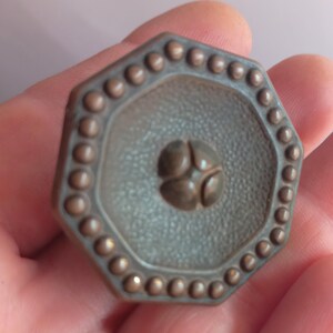 May include: An octagonal metal knob with a dark bronze finish. The surface features a textured, pebbled center with a raised four-petal floral motif in the middle. The outer edge is decorated with a continuous border of small, raised circular beads.