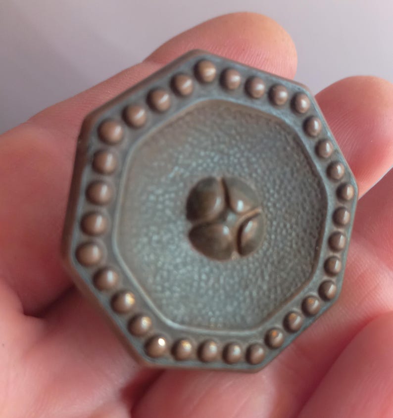 May include: An octagonal metal knob with a dark bronze finish. The surface features a textured, pebbled center with a raised four-petal floral motif in the middle. The outer edge is decorated with a continuous border of small, raised circular beads.