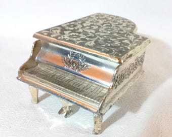 May include: A miniature silver-colored grand piano with intricate floral patterns on the lid. The piano features a detailed keyboard and decorative accents, with a small emblem on the front. The overall design is ornate and vintage.