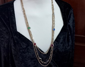 May include: A gold-toned necklace with multiple strands and colorful gemstone accents. The necklace features a variety of colored stones, including green, red, blue, and black. The necklace is displayed on a white mannequin against a black velvet background.