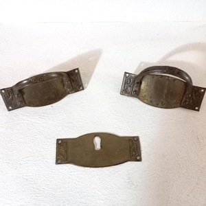 Puede incluir: Tres piezas de herrajes antiguos de latón. Dos son manijas con agarres curvos y bordes decorativos. La tercera es una placa de ojo de cerradura con un diseño a juego. Todas las piezas tienen un aspecto deslustrado y envejecido.