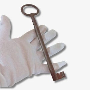 Puede incluir: Una llave larga de estilo antiguo con un asa circular y una paleta rectangular. La llave es de metal marrón óxido oscuro. La llave se sostiene en una mano enguantada sobre un fondo blanco.
