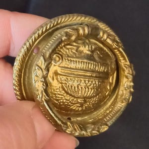 Puede incluir: Botón circular de latón dorado con un diseño ornamental. El botón presenta una imagen en relieve de un jarrón en el centro, rodeada por un borde detallado con elementos florales y de cuerda. Acabado envejecido.