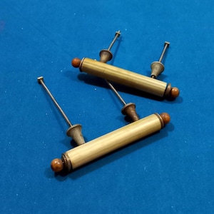 Puede incluir: Cuatro tiradores de cajón antiguos de latón con detalles de madera. Cada tirador presenta un cuerpo cilíndrico, un centro decorativo y un pequeño pomo de madera en cada extremo. Los tiradores están dispuestos sobre una superficie azul.