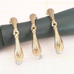 Op de afbeelding: Drie gouden kastdeurgrepen met een decoratief, geribbeld ontwerp. De grepen zijn bevestigd aan een donkergrijs oppervlak.