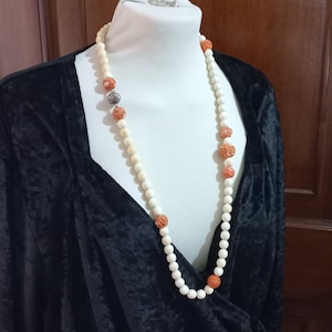 May include: A long necklace featuring cream-colored round beads interspersed with coral-colored, intricately carved beads. A silver-colored seashell charm is also included in the design. The necklace is displayed on a white mannequin.