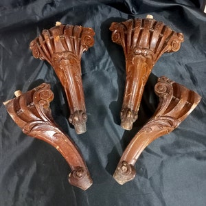 Könnte beinhalten: Vier antike Holzmöbelfüße mit aufwendigen Schnitzereien. Die Beine sind dunkelbraun mit detaillierten Schnörkeln und dekorativen Elementen. Die Beine stammen wahrscheinlich von einem Tisch oder Stuhl.