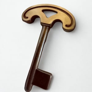 Puede incluir: Una llave decorativa con acabado bronce. La llave tiene una parte superior curvada con un diseño abierto y un vástago largo y rectangular. La cabeza de la llave tiene un diseño decorativo y abierto. La llave es un artículo decorativo de estilo vintage.