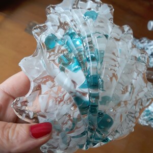 K&ouml;nnte beinhalten: Ein dekoratives Objekt in Form einer Muschel aus klarem Glas mit t&uuml;rkisfarbenen und wei&szlig;en Akzenten. Das Glas hat ein strukturiertes, rissiges Aussehen. Das Objekt wird in einer Hand mit rotem Nagellack gehalten.