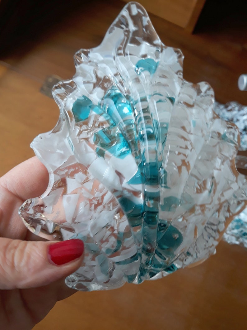 K&ouml;nnte beinhalten: Ein dekoratives Objekt in Form einer Muschel aus klarem Glas mit t&uuml;rkisfarbenen und wei&szlig;en Akzenten. Das Glas hat ein strukturiertes, rissiges Aussehen. Das Objekt wird in einer Hand mit rotem Nagellack gehalten.