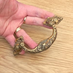 Könnte beinhalten: Verzierter Schubladengriff aus Messing mit gebogenem Griff und dekorativen floralen und scrollartigen Details. Die Hardware hat eine Vintage-Ästhetik mit einer leicht gealterten Patina. Für Möbel und Schränke.