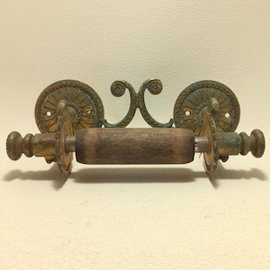 Puede incluir: Un portarrollos de papel higiénico de estilo antiguo con un rodillo de madera. El soporte presenta monturas circulares de latón ornamentadas con detalles florales y un acento decorativo de latón. El latón tiene una apariencia desgastada y envejecida, lo que aumenta su encanto vintage.