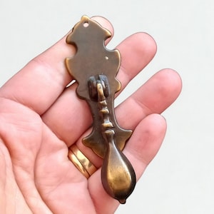 May include: Antique brass drawer pull with a decorative backplate and a teardrop-shaped handle. The pull has a vintage aesthetic, with a tarnished finish and intricate detailing. The hardware is designed for furniture restoration or adding a classic touch to cabinets.
