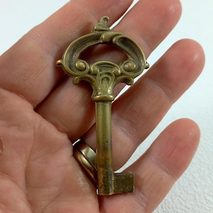 May include: A vintage brass key with an ornate, decorative head. The key has a long, thin shaft and a square-shaped end.