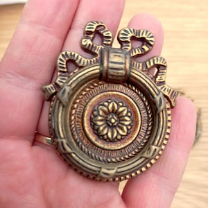 Puede incluir: Tirador de cajón de latón antiguo con un diseño decorativo de lazo y anillo. El centro presenta un motivo floral dentro de un marco circular. El tirador tiene una estética vintage, adecuado para la restauración de muebles.
