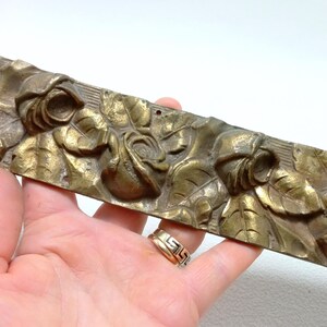 Puede incluir: Una moldura decorativa de latón vintage con un relieve de rosas y hojas. La moldura tiene forma rectangular y un aspecto envejecido y deslustrado, con detalles del diseño floral. La pieza se sostiene en una mano.