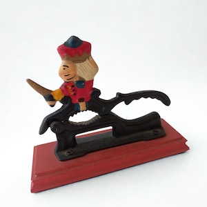 Pode incluir: Um quebra-nozes vintage com uma figura de soldado sobre um mecanismo de metal preto, montado em uma base de madeira vermelha. O soldado usa uma jaqueta vermelha, um chapéu azul e cabelo loiro. O quebra-nozes é projetado para quebrar nozes.