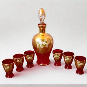 Könnte beinhalten: Ein Vintage-Dekanter-Set aus rotem und goldenem Glas. Die Karaffe hat einen goldenen Körper mit rotem Sockel und einen durchsichtigen Stopfen. Sechs passende rote Gläser mit goldenen Rändern und Blumendesigns sind enthalten.