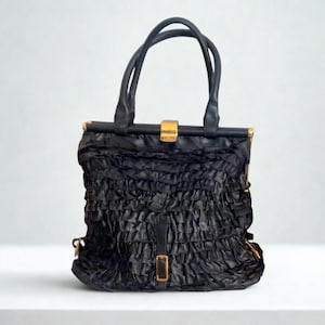 May include: A white ceramic mug with a black handle and the text "But First, Coffee" in a bold, black font. The mug is set against a blurred background. The mug appears to be approximately 10 cm tall.