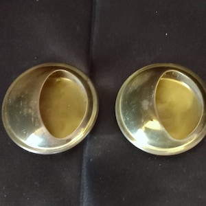 Puede incluir: Dos tiradores de armario circulares dorados, cada uno con una hendidura en forma de media luna. Los tiradores tienen una superficie lisa y reflectante, sobre un fondo oscuro. El diseño es simple y moderno, adecuado para varios estilos de muebles.