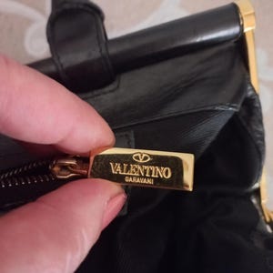 May include: Black leather handbag with gold-tone detailing. The bag has a top handle and a rectangular shape. The Valentino Garavani logo is on a gold metal plate.