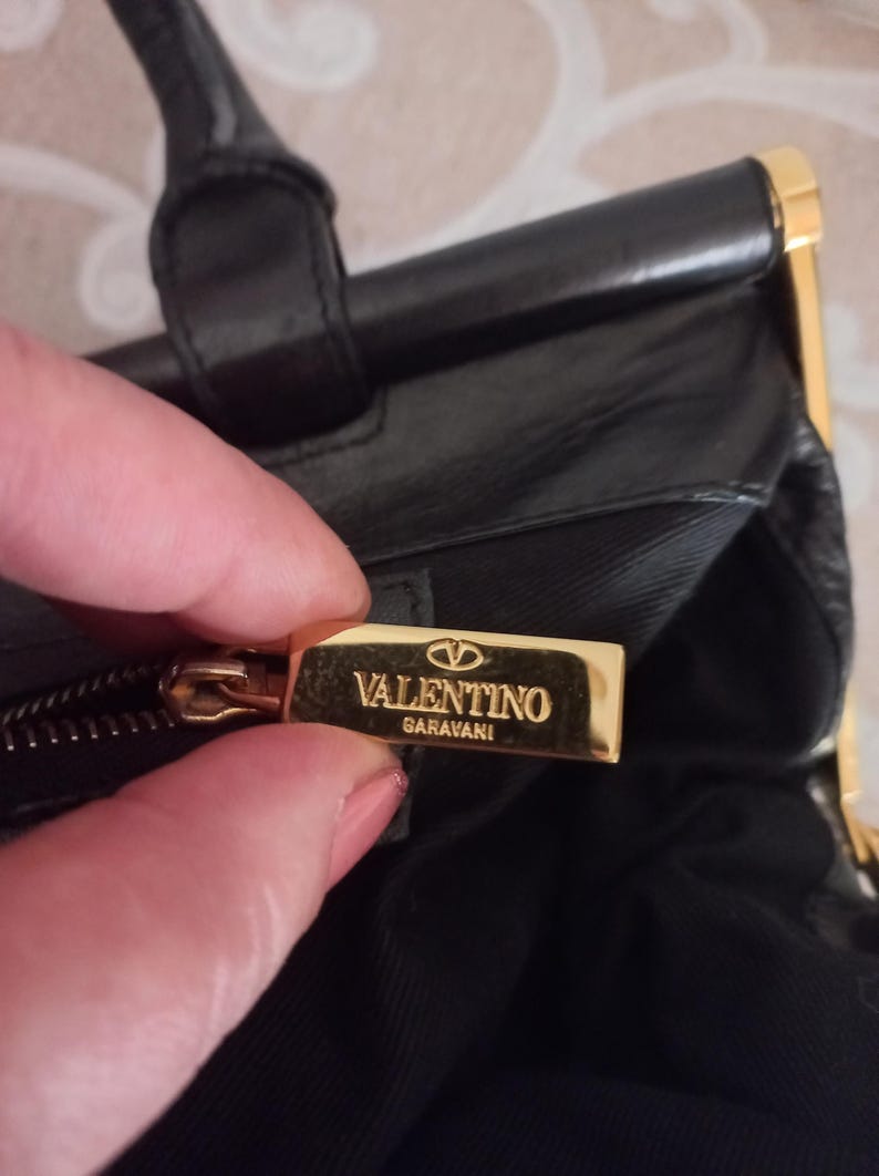 May include: Black leather handbag with gold-tone detailing. The bag has a top handle and a rectangular shape. The Valentino Garavani logo is on a gold metal plate.