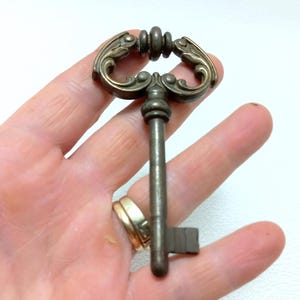 以下が含まれることがあります： 装飾的で装飾的なハンドルが付いたアンティークスタイルのキー。キーはブロンズ色の金属でできており、複雑なスクロールワークと詳細なボウが特徴です。キーは手で持たれており、そのサイズとデザインを示しています。