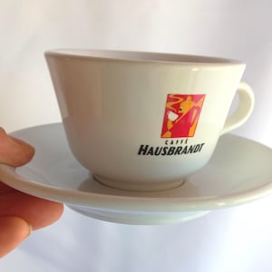 May include: White ceramic coffee cup and saucer. The cup has a handle and a logo with red, orange, and yellow accents, and the text "CAFFE HAUSBRANDT". The saucer is round and white.