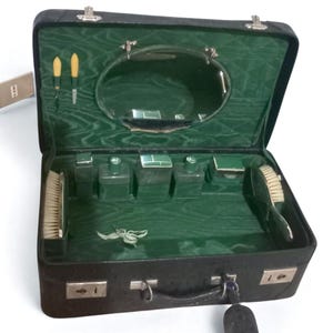 Puede incluir: Maleta de aseo de viaje vintage negra abierta con interior de terciopelo verde. El kit incluye un espejo, cepillos, botellas y herramientas de manicura. La maleta es rectangular con herrajes plateados y un asa.