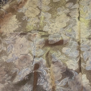 Puede incluir: Primer plano de una tela floral con fondo dorado metálico. La tela presenta un patrón repetido de grandes flores y hojas de color azul claro y blanco, creando una apariencia texturizada. Adecuado para ropa o decoración del hogar.