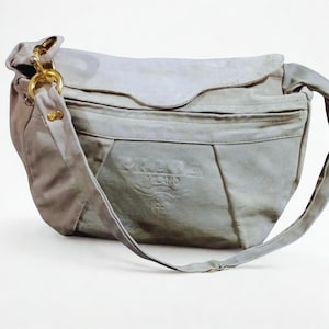 May include: A light gray Prada shoulder bag with a flap closure and a long strap. The bag features gold-tone hardware and the Prada logo embossed on the front. The bag is made of a soft, suede-like material.