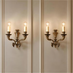 Puede incluir: Un par de apliques de pared de dos brazos hechos de metal con tono de latón antiguo. Cada lámpara presenta un remate decorativo central, dos brazos curvos con arandelas en forma de hoja y dos bombillas verticales estilo vela.