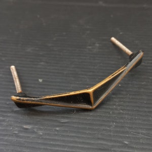 Könnte beinhalten: Ein schwarzer und goldener Metall-Schranktürgriff mit geometrischem Design. Der Griff hat an beiden Enden zwei Schrauben.