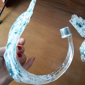 K&ouml;nnte beinhalten: Ein dekoratives Objekt aus klarem Glas mit einem geschwungenen, wellenartigen Design. Das Glas hat ein mattiertes Aussehen mit blauen und wei&szlig;en Streifen. Eine silberne Metallkappe ist am Ende des gebogenen Glases befestigt.