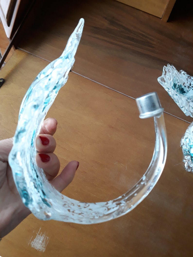 K&ouml;nnte beinhalten: Ein dekoratives Objekt aus klarem Glas mit einem geschwungenen, wellenartigen Design. Das Glas hat ein mattiertes Aussehen mit blauen und wei&szlig;en Streifen. Eine silberne Metallkappe ist am Ende des gebogenen Glases befestigt.