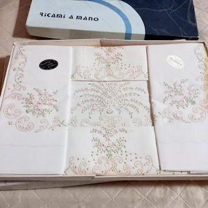 Può includere: Un set di biancheria ricamata bianca, tra cui tovaglioli e un pezzo più grande, presentato in una scatola. Il ricamo presenta delicati disegni floreali in rosa e verde. Il coperchio della scatola riporta la scritta "Ricami a mano".