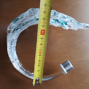 K&ouml;nnte beinhalten: Eine klare Glasskulptur mit wellenartigem Design, akzentuiert mit T&uuml;rkis. Die Skulptur ist etwa 15 cm hoch. Sie hat einen silbernen Metallsockel. Das St&uuml;ck befindet sich auf einer braunen Oberfl&auml;che.