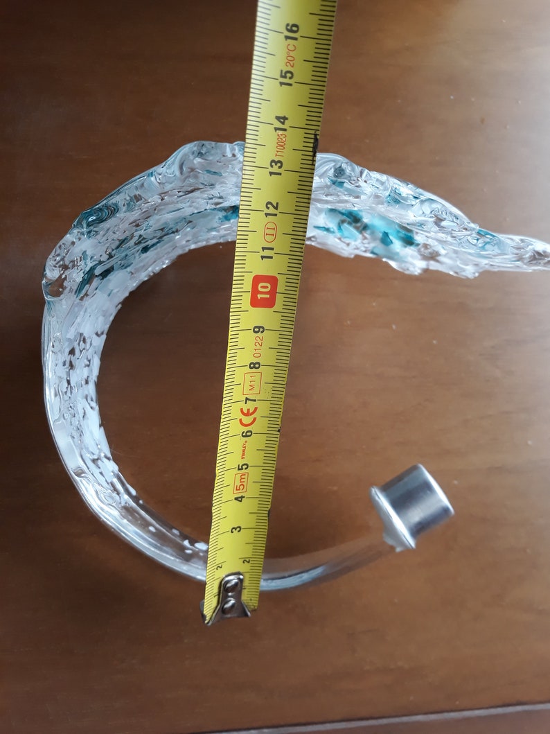 K&ouml;nnte beinhalten: Eine klare Glasskulptur mit wellenartigem Design, akzentuiert mit T&uuml;rkis. Die Skulptur ist etwa 15 cm hoch. Sie hat einen silbernen Metallsockel. Das St&uuml;ck befindet sich auf einer braunen Oberfl&auml;che.