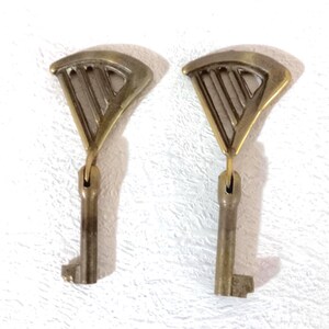 Puede incluir: Dos llaves de latón de estilo antiguo. Cada llave tiene una parte superior en forma de abanico con un diseño geométrico y una paleta rectangular. Las llaves están suspendidas de un pequeño lazo.